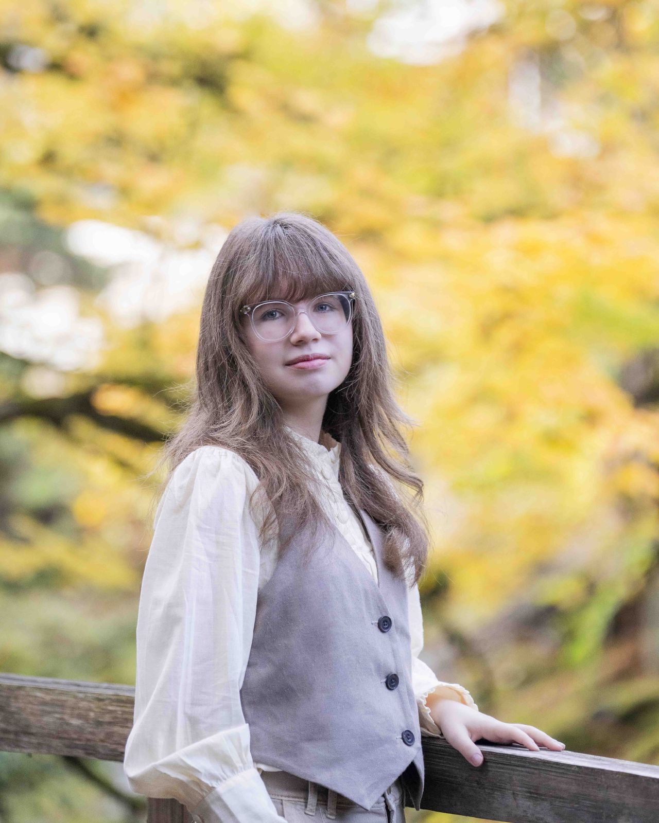 A young woman in a vest and wide-leg trousers leans on a fence by a creek, framed by golden autumn leaves
