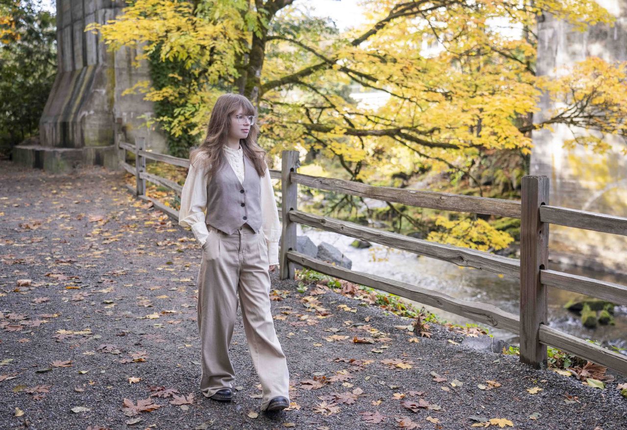 A young woman in a vest and wide-leg trousers walks along a path near a creek, framed by golden autumn leaves.