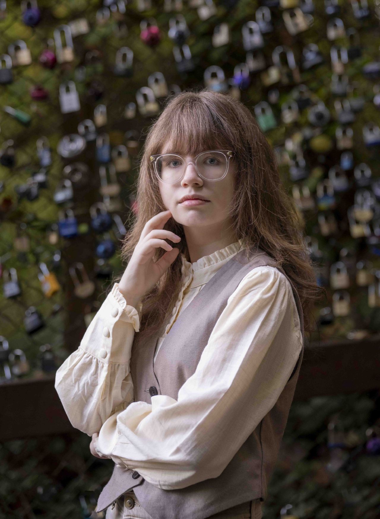 A young woman in a vest frames her face with her hand, in front of a fence covered in padlocks