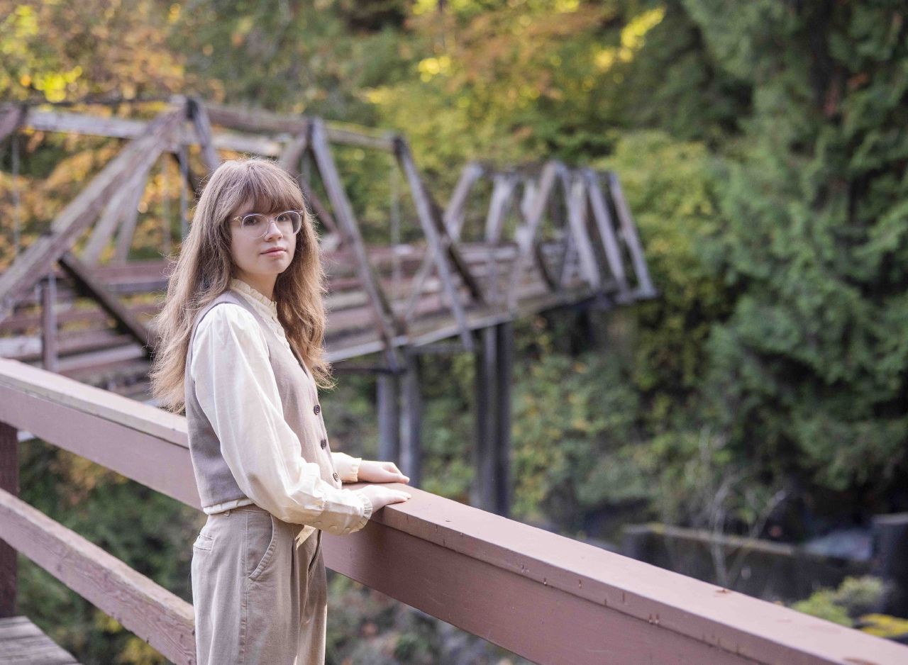A young woman in a vest and wide-leg trousers leans on a fence by a creek, framed by golden autumn leaves and a wooden bridge.