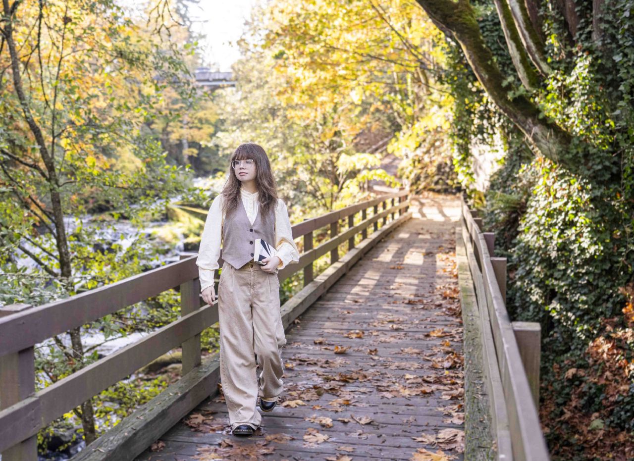 A young woman in a vest and wide-leg trousers holding a book near a fence by a creek, framed by golden autumn leaves and a wooden bridge