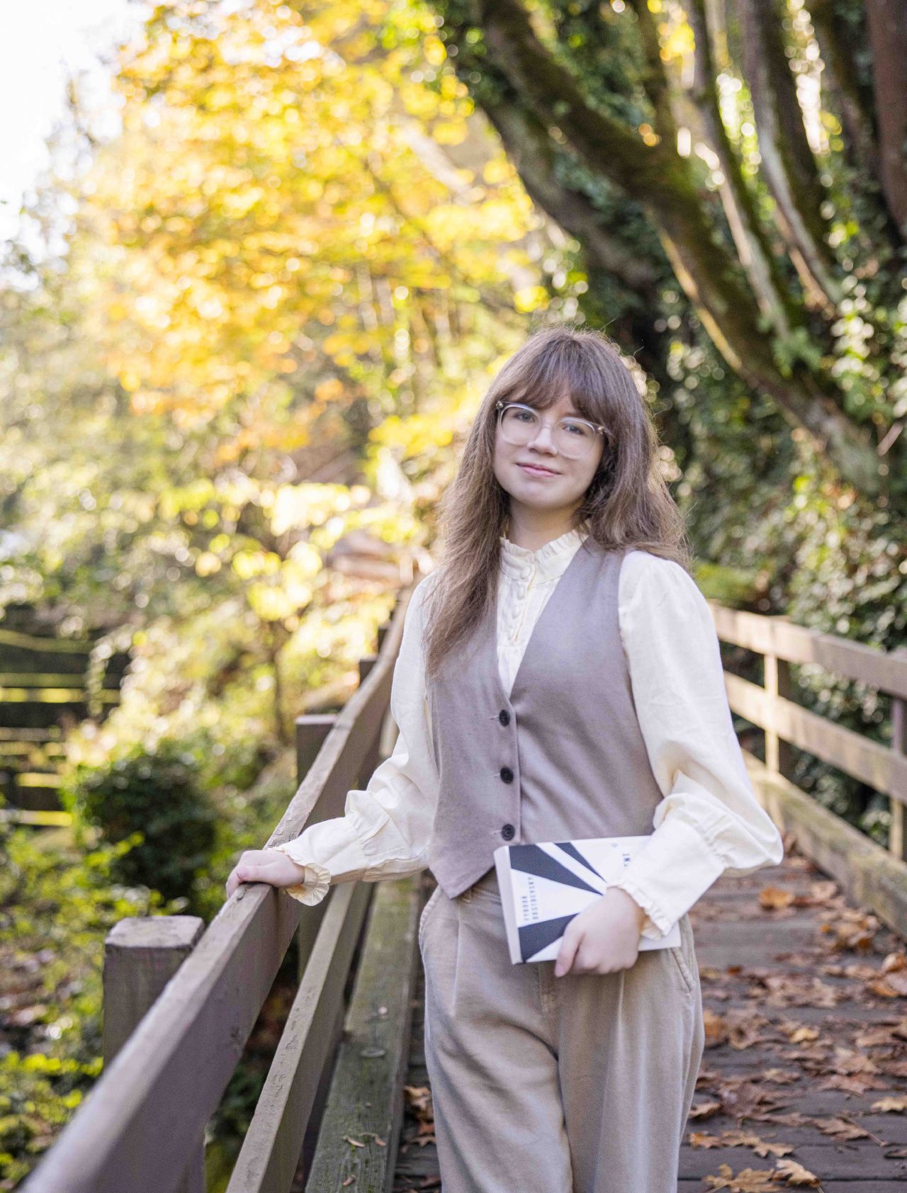 A young woman in a cream colored blouse with a vest and round glasses, holding a book and a fence, looks softly at the camera on an autumn day.