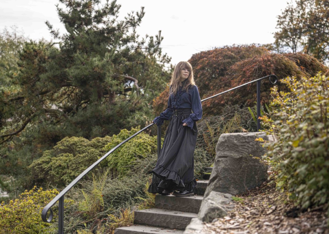 A woman in a vintage blue blouse and black maxi skirt walks up a stone staircase in a lush garden, holding the handrail and gazing ahead.