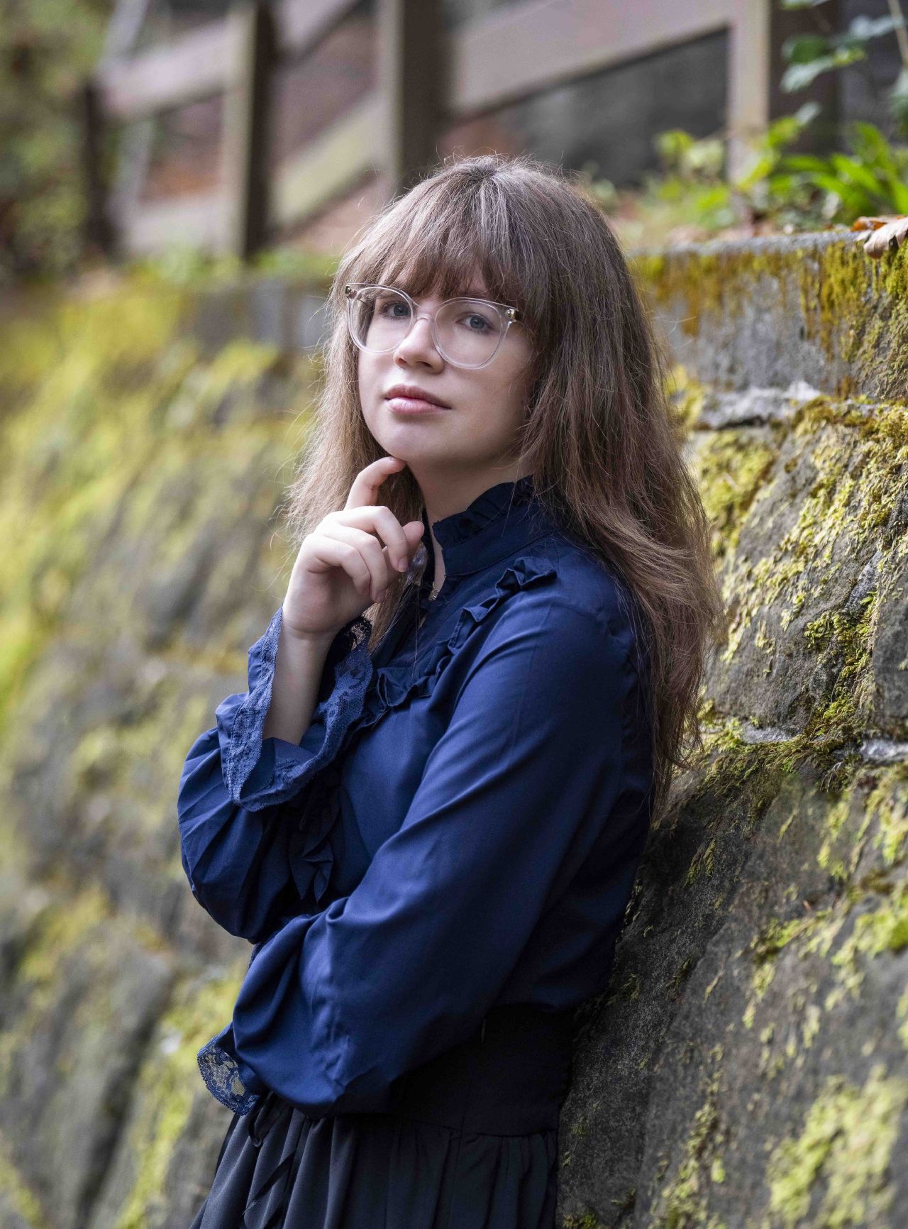 A young woman in a vintage blue blouse with round glasses looks confidently directly at the camera while leaning against a moss covered wall