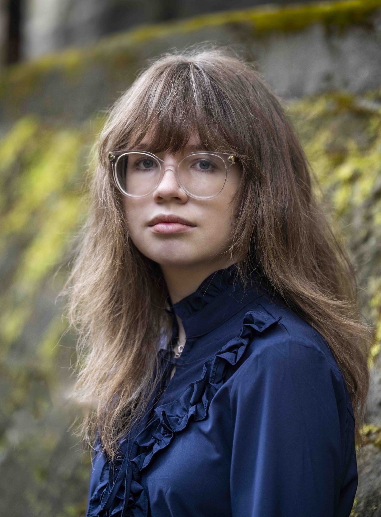 A young woman in a vintage blue blouse with round glasses looks confidently directly at the camera