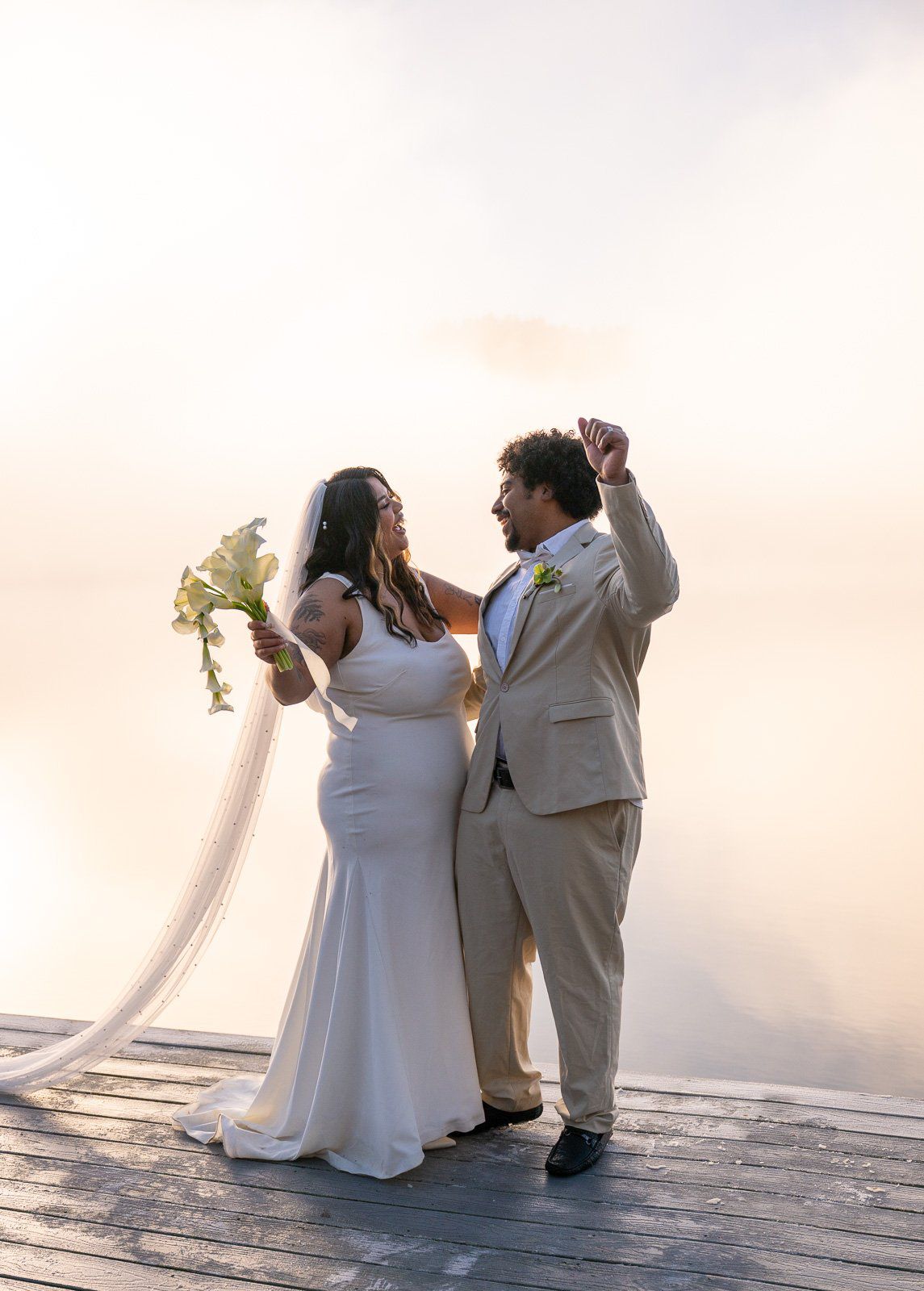 Bride and groom laughing together on the dock, groom's hand raised mid-twirl, morning fog rising off the lake behind them.