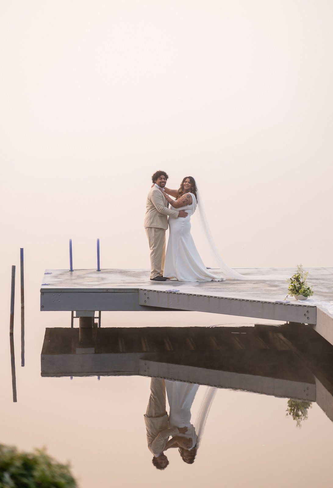 Bride and groom standing close together at the end of the dock, embracing and looking toward the camera, their reflection visible below.