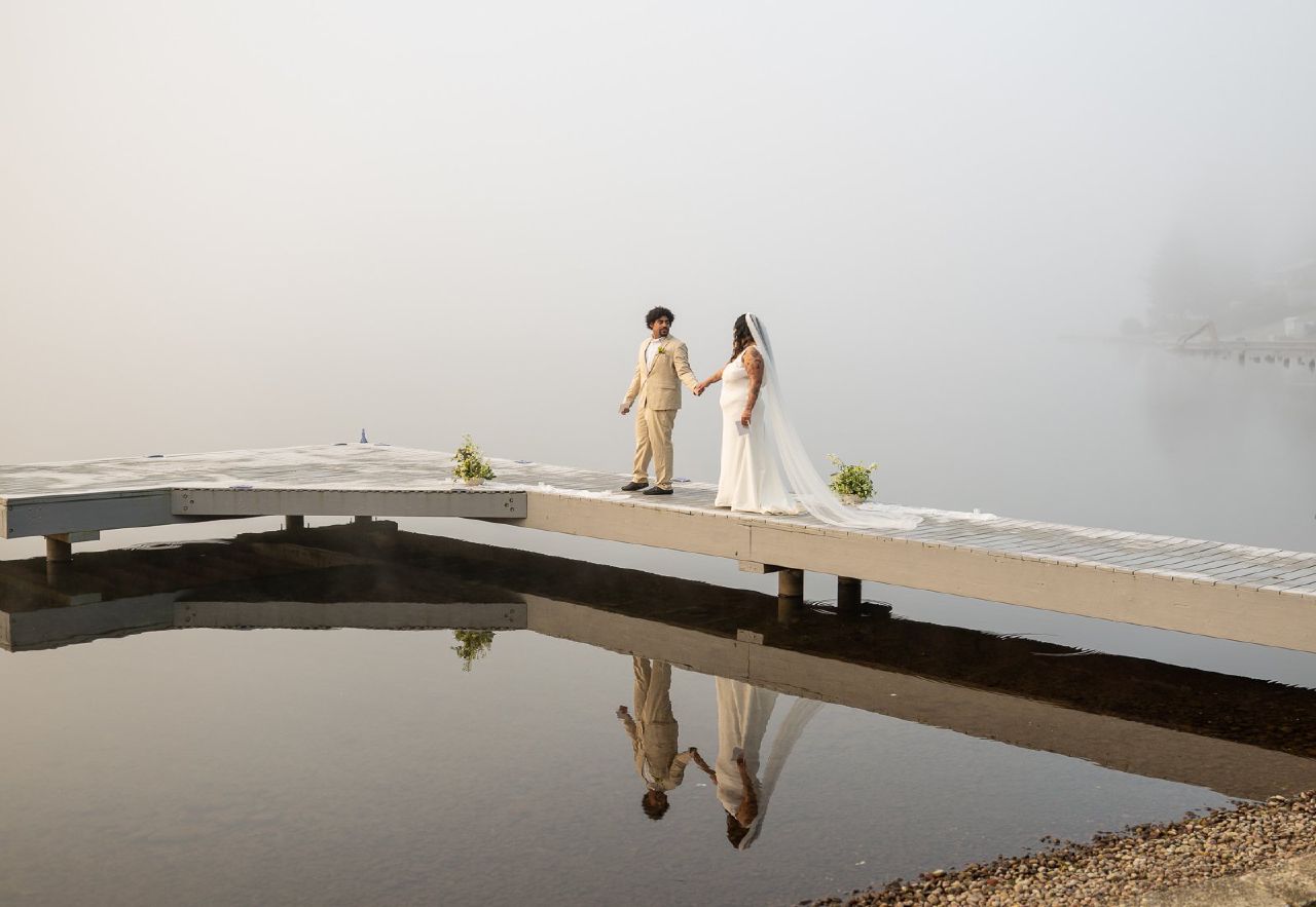 Groom glancing back at the bride as they walk down the dock, her veil trailing behind them through the fog and marina.
