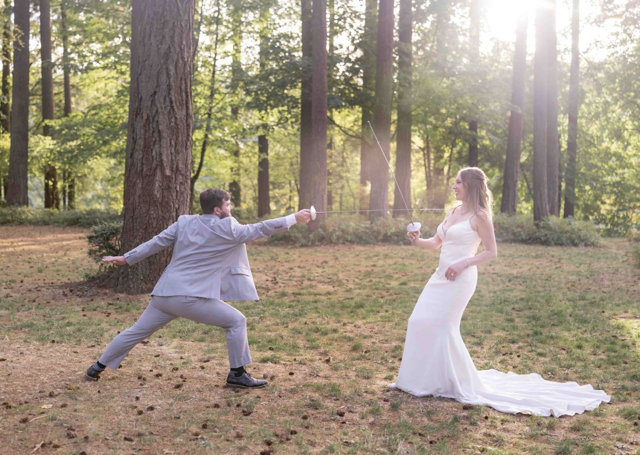 Groom playfully lunges with a fencing sword toward bride in the woods at Lacamas Lake Park.