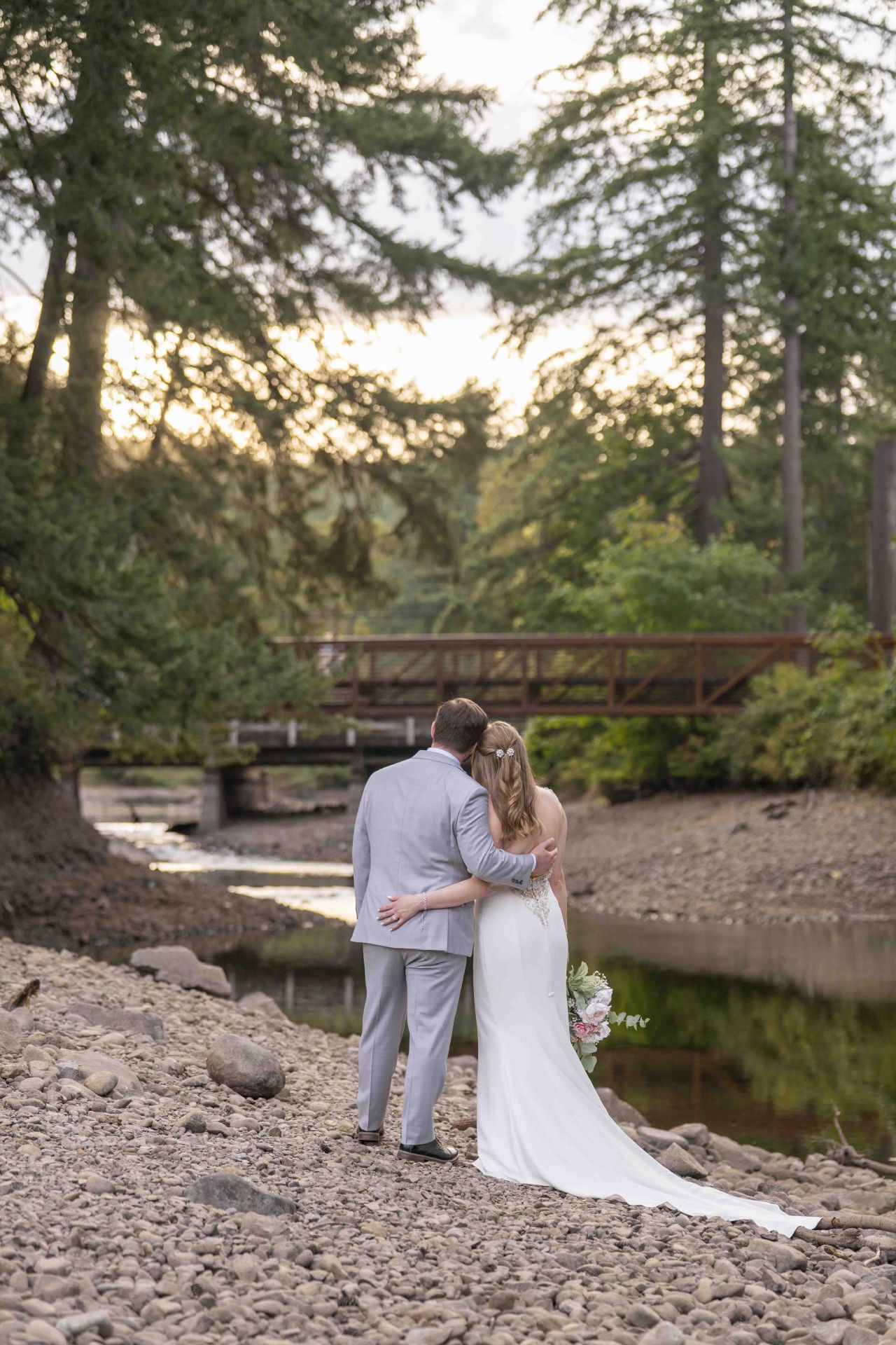 Bride and groom embrace by the creek and bridge at Lacamas Lake Park in Camas, Washington.