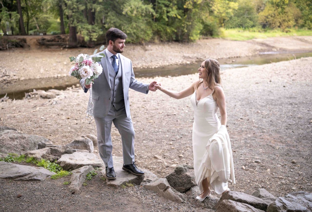 Groom helps bride step over rocks by the creek at Lacamas Lake Park, holding her bouquet of blush flowers.